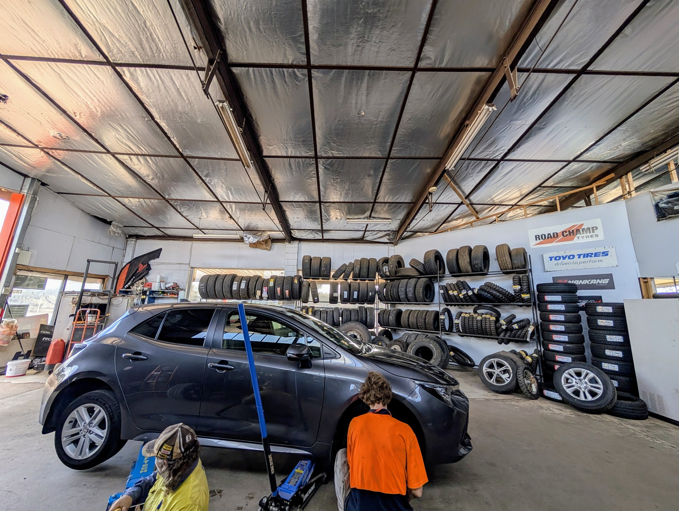 Two staff working on a car in the Steel City Tyres workshop with racked tyres and stacked alloy wheels behind